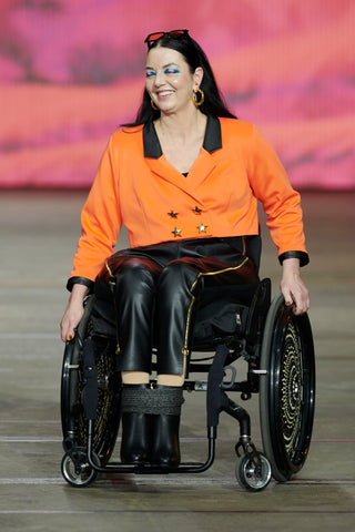 A dark haired woman in a wheelchair wearing an orange and black jacket and black leather pants