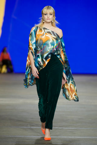 A blonde haired woman wearing a silk patterned off one shoulder top and high waisted emerald green pants with bright orange heels 