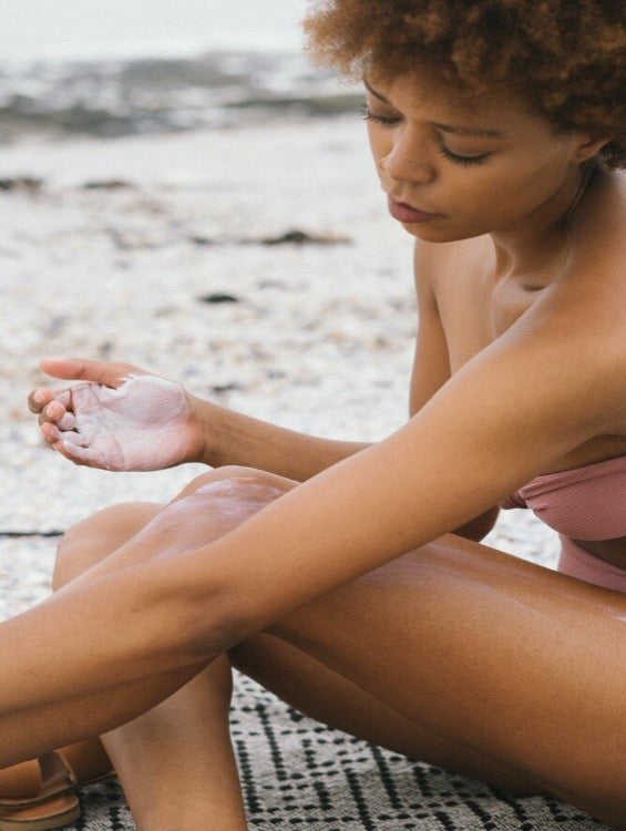 Woman with dark skin in a bikini sits on a towel on the beach and puts on lotion