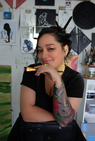 An artist straddles her rolling studio chair confidently in her studio, adorned with a beautiful tattoo, surrounded by a vibrant array of her stunning artworks. The room is filled with creativity, showcasing the artist's talent and the captivating beauty of her diverse portfolio