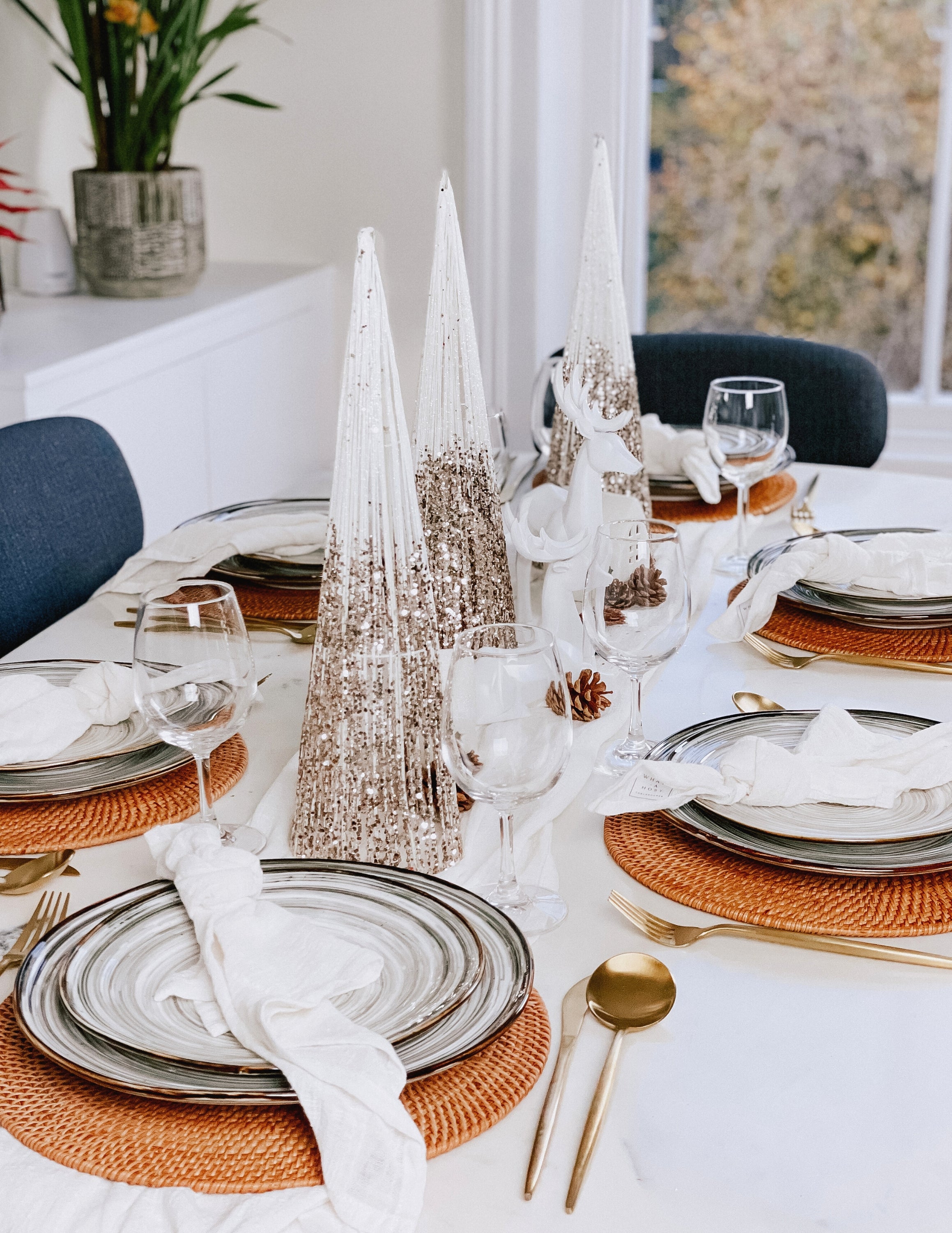 What a Host Home Decor: Christmas Tablescape With Geometric Decorative Christmas Deers, Scandi Houses, Rattan Placemats, Porcelain Plates Set Tableware, White and Gold Christmas Table Pine Cones, Gold Cutlery Stainless Steel Set and Rustic Cotton Gauze Table Textiles