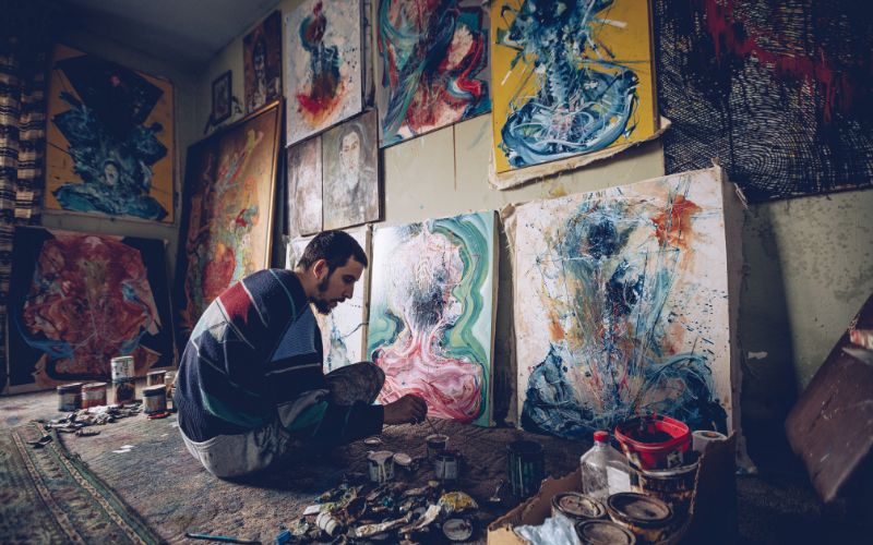 An artist sitting on the floor while painting on canvas