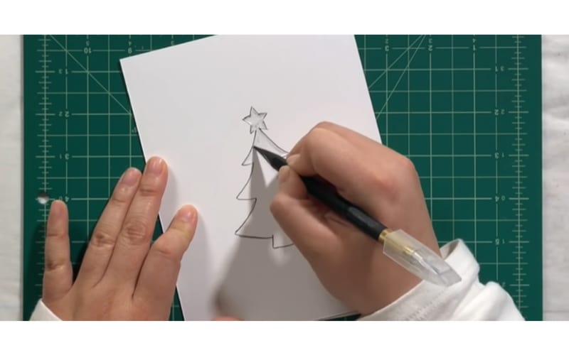 a crafter embossing a Christmas tree on thick white paper using a stencil as a guide