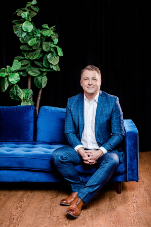 Professional portrait of a man in a blue suit sitting on a couch. The man exudes confidence and professionalism in this posed headshot