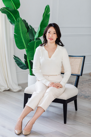 Professional headshot of a confident woman of Asian descent, dressed in white, sitting in a chair and posing with professionalism.
