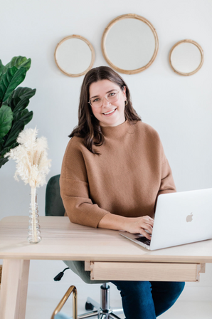 Professional headshot of a smiling woman sitting in a chair with a laptop on the table in front of her. The woman exudes confidence and professionalism in her pose