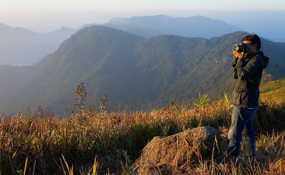 A man taking a picture on a hill with mountains in the background