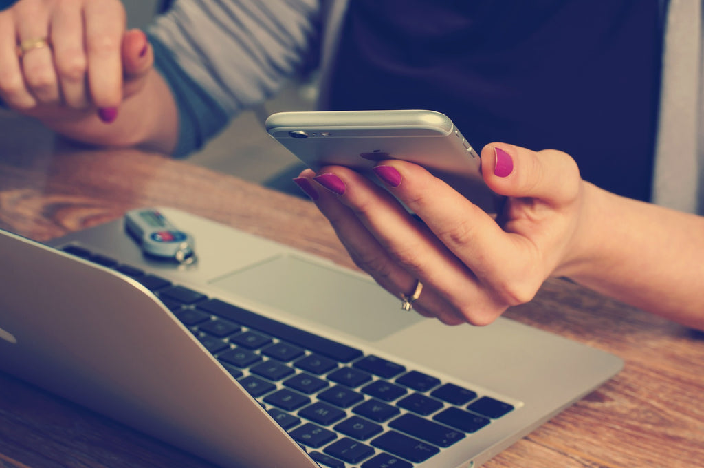 Close up of woman's hand looking at phone while sat at a table with a laptop.