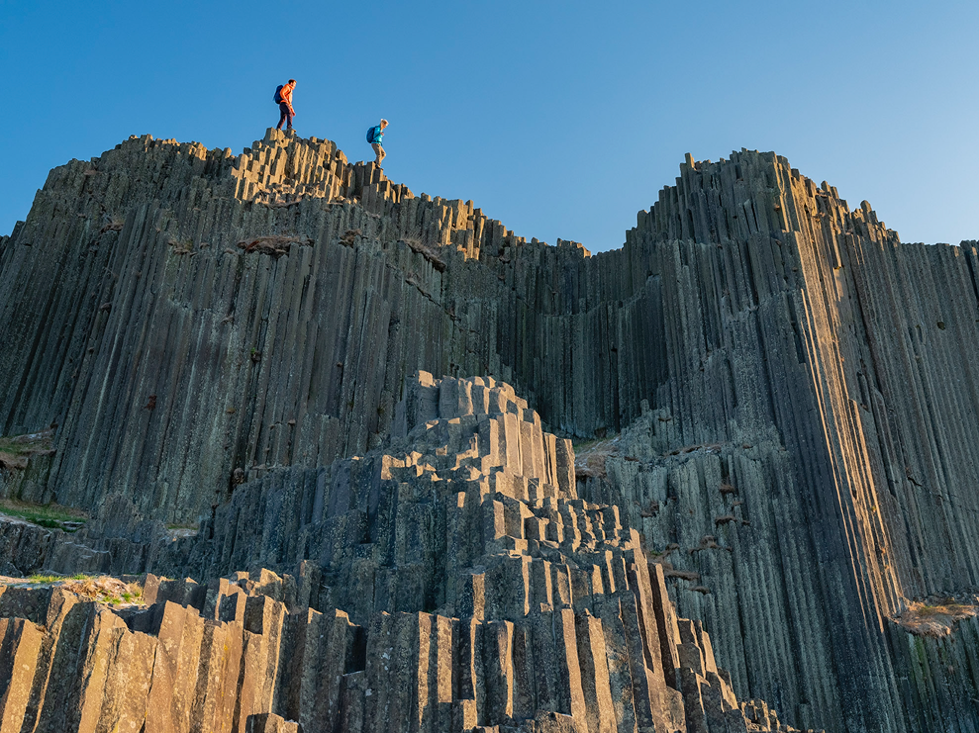 Two walkers stood on top of a rock with the sun shining. 