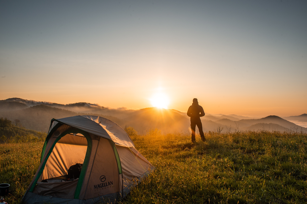 A hiker on a hill looking at the sunset with his tent on an overnight hike