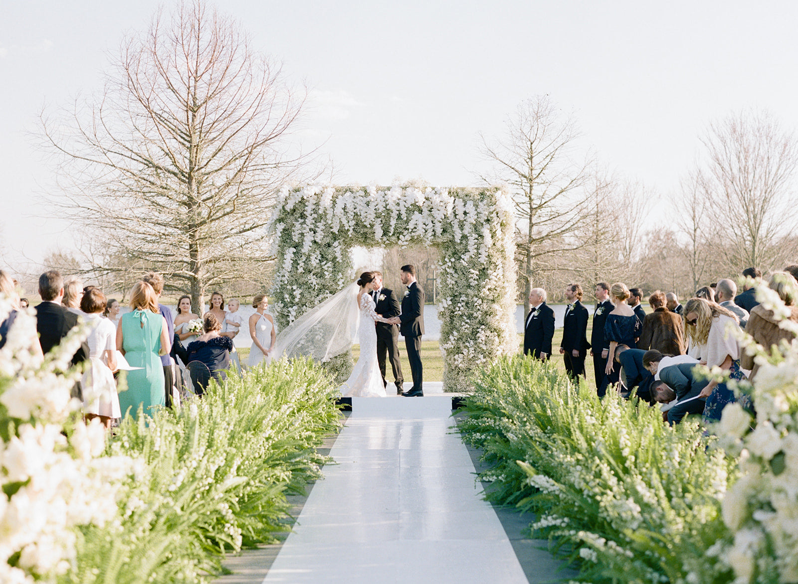 Photo of a bridge and groom standing beneath a square arch made of greenery and flowers in front of a large audience of wedding guests