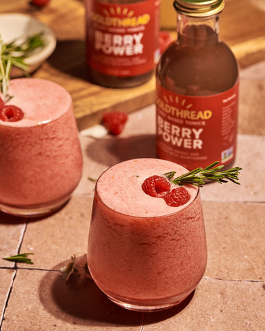 Goldthread Plant-Based Tonics Frozen Boozy Berry Lemonade garnished with raspberries and rosemary next to Goldthread Plant-Based Tonics Berry Power tonic bottles