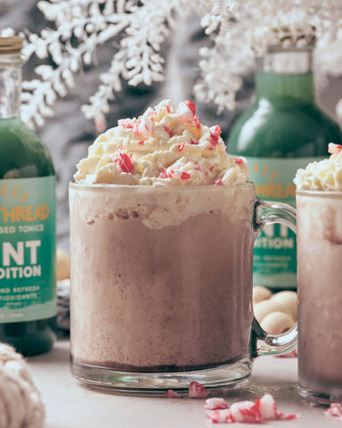 Goldthread Plant-Based Tonics Frozen Mint Hot Chocolate garnished with crushed candy canes and whipped cream with Goldthread Plant-Based Tonics Mint Condition tonic bottles