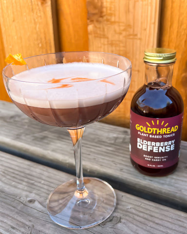 Goldthread Tonics Elderberry Whiskey Sour Cocktail in coupe glass sitting on wooden bench next to Goldthread Tonics Elderberry Defense Tonic