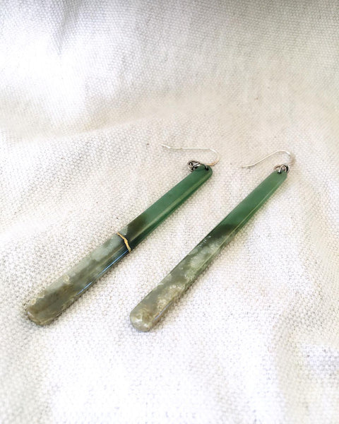 A pair of long, slender greenstone (pounamu) earrings on a white textured cloth; one was broken in two pieces and has been repaired using the traditional kintsugi technique, evident by the gold seam binding the break.