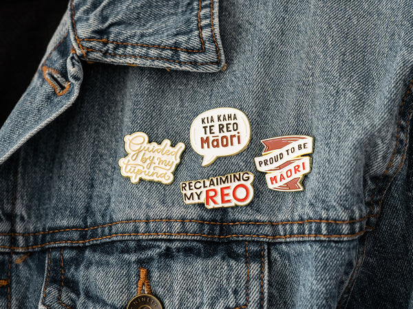 A close-up photo of a denim jacket with four enamel pins attached. From left to right, the pins read: "Guided by my tūpuna," "Kia kaha te reo Māori," "Reclaimin my Reo," and "Proud to be Māori." The pins are designed with decorative elements and colors to emphasize the messages of guidance by ancestors, strength for the Māori language, and Māori pride.