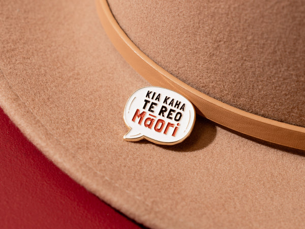 An enamel badge pin with the words "Kia Kaha Te Reo Māori" written in bold letters on a tan wool hat