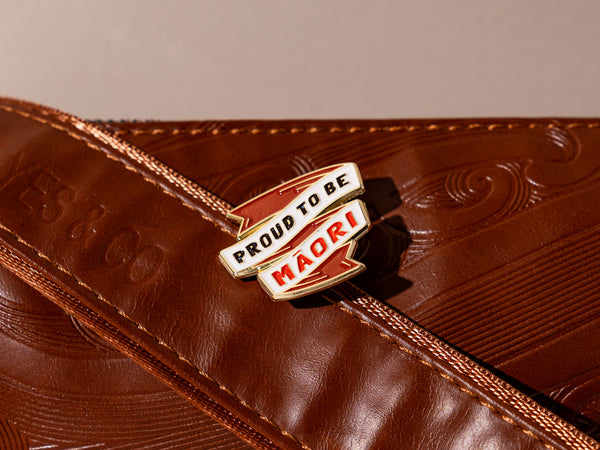 An enamel badge pin with the words "Proud to be Māori" written in bold letters on a brown leather bag with Māori motifs and designs