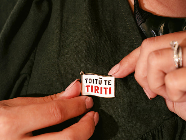 Two hands securing an enamel pin in the shape of a flag with the words "Toitū te Tiriti" written in bold letters onto a dark forest green dress