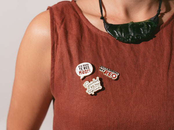 A person is wearing a clay colour dress adorned with three enamel pins, featuring the phrases "Kia kaha te reo Māori", "Guided by my tūpuna" and "Reclaiming my REO." Above the pins, the person is wearing a traditional Māori carved pounamu (greenstone) necklace.