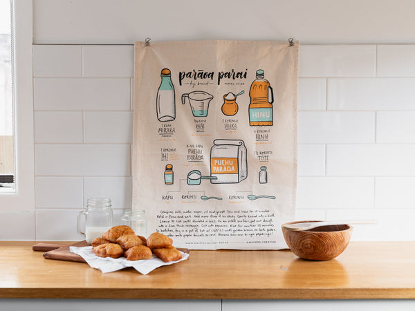 A kitchen setting with a tea towel hung on the wall featuring illustrations and recipe instructions for making frybread, accompanied by a plate of freshly made frybread on a wooden board, a mason jar, and a wooden bowl on a countertop.