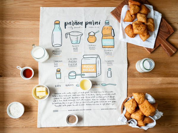 Overhead view of a table with a tea towel illustrated with a fry bread recipe and drawings of ingredients. Around the tea towel are the actual ingredients including oil, flour, yeast, sugar, a jar of milk, butter, and two stacks of fry bread on wooden boards.