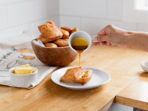 A person's hand pouring golden syrup over a piece of Māori fry bread on a white plate, with a bowl of additional fry bread and a dish of butter cubes in the background, set on a wooden kitchen counter.