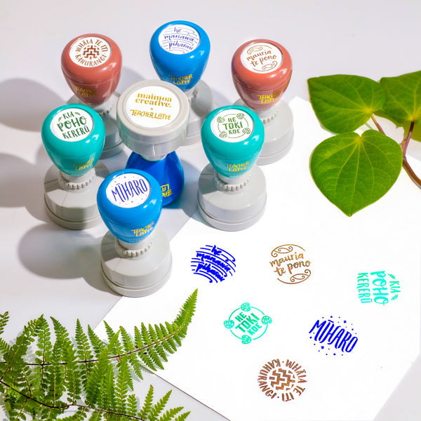 An array of colourful self-inking teacher stamps with phrases in te reo Māori stamped below them, surrounded by vibrant greenery on a white surface