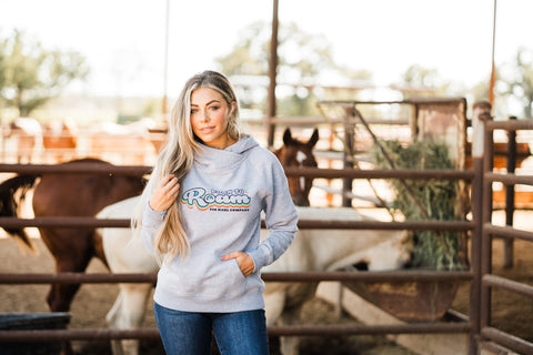 A fashion-forward woman in a 'Born to Roam' Tin Haul hoodie stands confidently at a ranch with horses in the background, symbolizing the modern twist on Western wear for 2024. Her casual yet chic ensemble pairs the cozy sweatshirt with classic blue jeans, encapsulating the blend of comfort and style that defines the latest trends in country clothing.