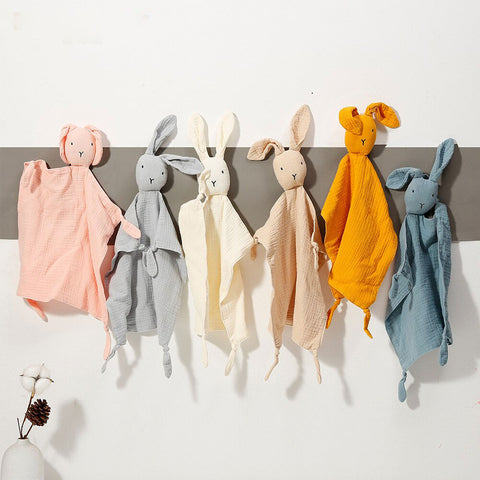 Six doudous doux de couleurs (rose, bleu, blanc, beige, moutarde et gris) suspendus sur un porte-manteau, créant une ambiance chaleureuse et réconfortante. Idéal pour un cadeau de naissance.