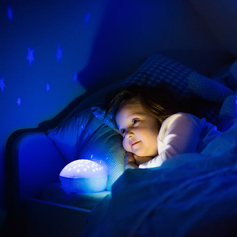 Une petite fille émerveillée dans son lit, regardant les étoiles projetées sur le mur de sa chambre par une veilleuse, illustrant l'effet apaisant et fascinant des veilleuses qui projettent des images, aidant les enfants à s'endormir tout en stimulant leur imagination.