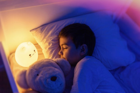Un enfant paisiblement endormi dans son lit, éclairé par la douce lueur d'une veilleuse allumée, avec une peluche confortablement nichée à côté de lui, illustrant l'efficacité des veilleuses pour aider les enfants à s'endormir.