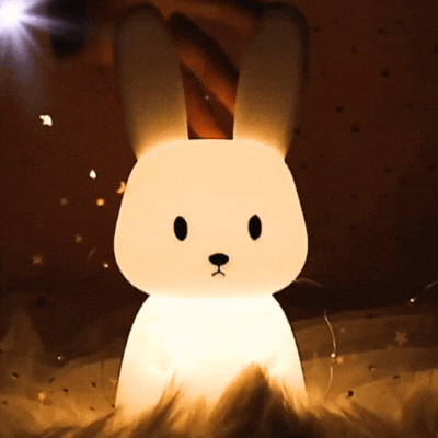 Main touchant une veilleuse lapin en silicone souple, illuminée dans un environnement féerique.