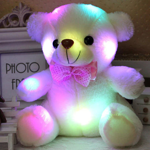 Une veilleuse peluche en forme d'ourson blanc avec un nœud papillon rose, illuminée de plusieurs couleurs, illustrant une option de veilleuse douce et réconfortante pour les enfants qui combine la chaleur d'une peluche avec la fonctionnalité d'une veilleuse multicolore.