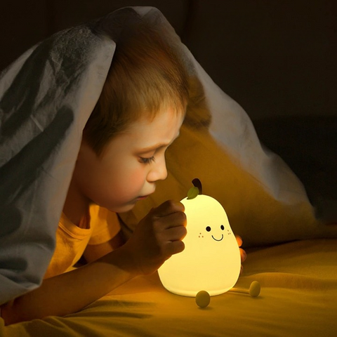 Un enfant dans son lit sous sa couette, touchant doucement une veilleuse en forme de poire illuminée d'une couleur chaude, illustrant l'interaction des enfants avec les veilleuses tactiles et l'atmosphère apaisante qu'elles créent pour favoriser un sommeil paisible.