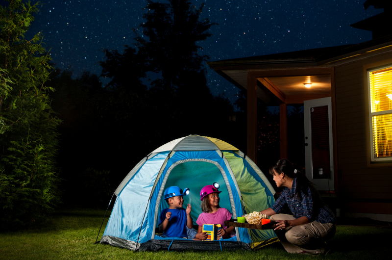 two young kids with headlamps on in a tent in a backyard at night with stars in the sky and a mom bringing them a snack of popcorn