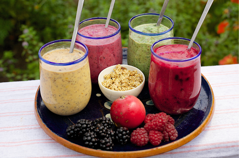 four glasses with straws of different flavors of smoothies on a tray with fresh berries and granola