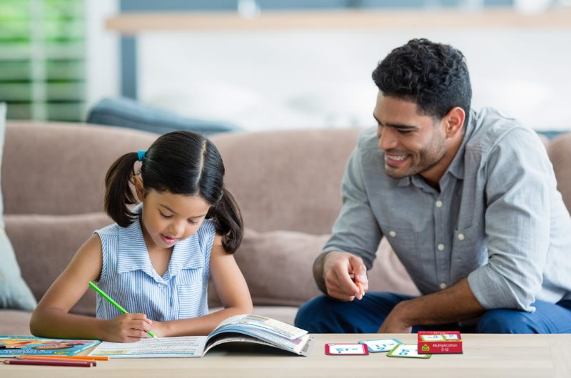 dad sitting on a couch while his daughter is sitting at the coffee table working in a math workbook