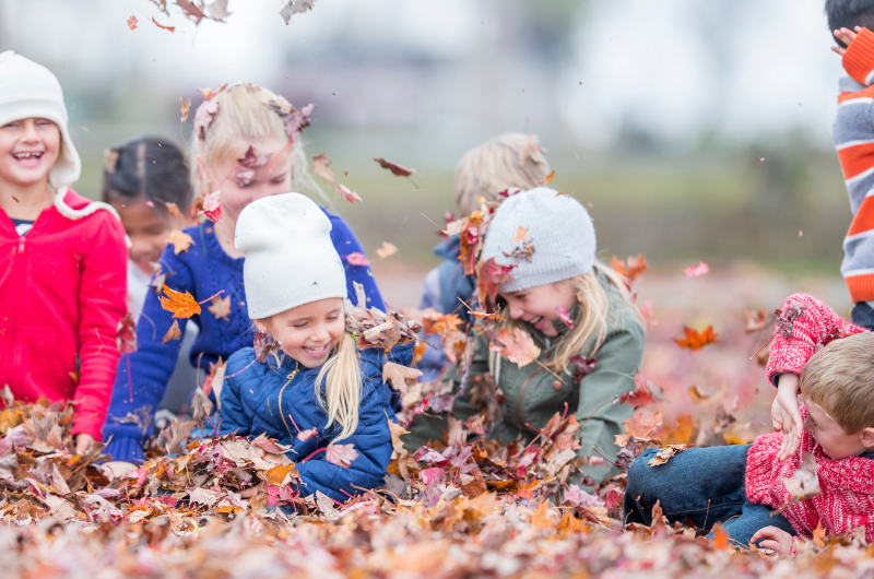 a fall scene of kids in coats and hats laughing and playing in fallen leaves