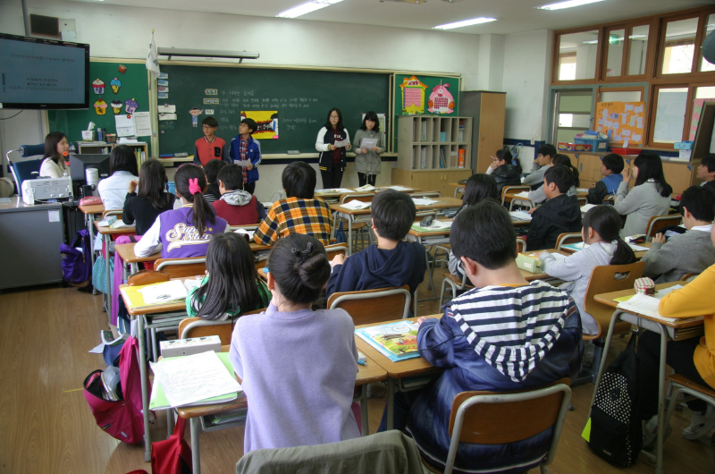 photo from the back of a full school classroom of 3-4th grade kids at their desks with students standing at the front giving reports