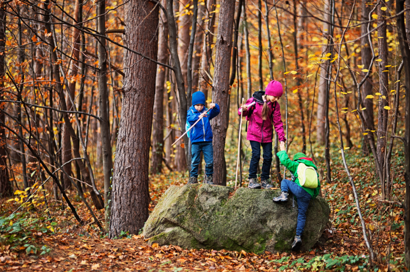 three kids on a fall hick climbing on a large boulder with trees that have changed color in the background behind them