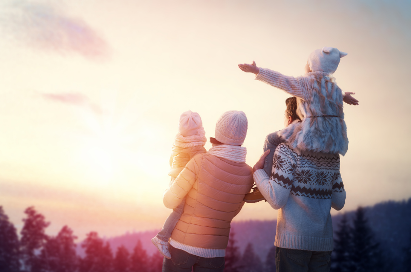 family in winter mon holding one kid and dad with a kid on his shoulders bundled up outside excited as they face the sun to watch the sunrise