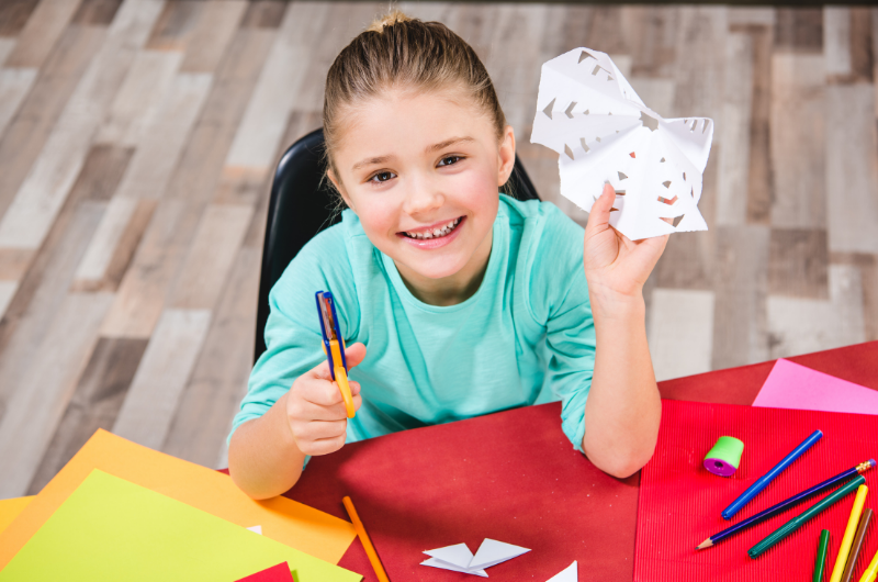 young girl in a classroom holding up a paper snowflake she just made and smiling