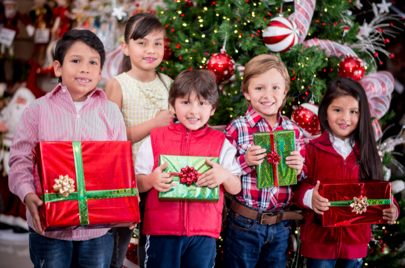 group of five young kids in front of a Christmas tree holding gifts