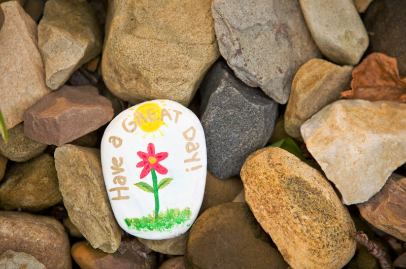 a painted rock with the message Have A Good Day and a red flower in the middle laying amongst many unpainted rocks