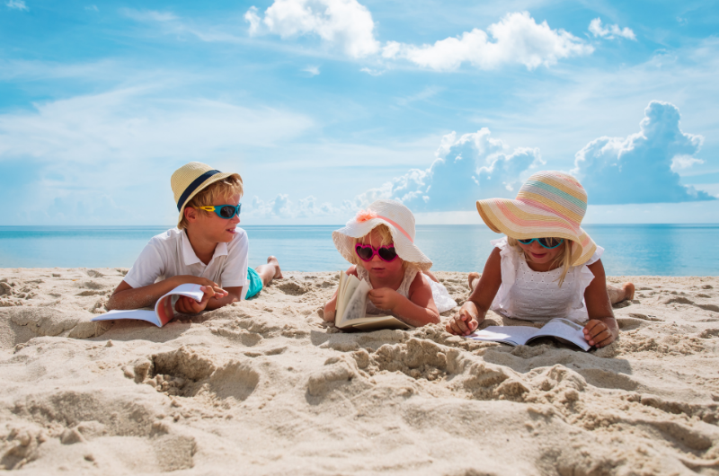 three kids on a beach laying in the sand with sunglasses on and sun hats reading books