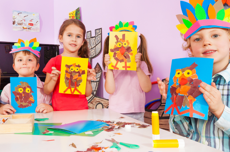 four kids standing around a craft table with construction paper making art projects holding up pictures of torn paper owls