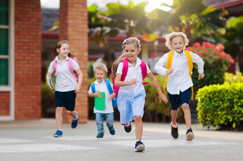 four elementary age kids with backpacks on running along the sidewalk by a school 