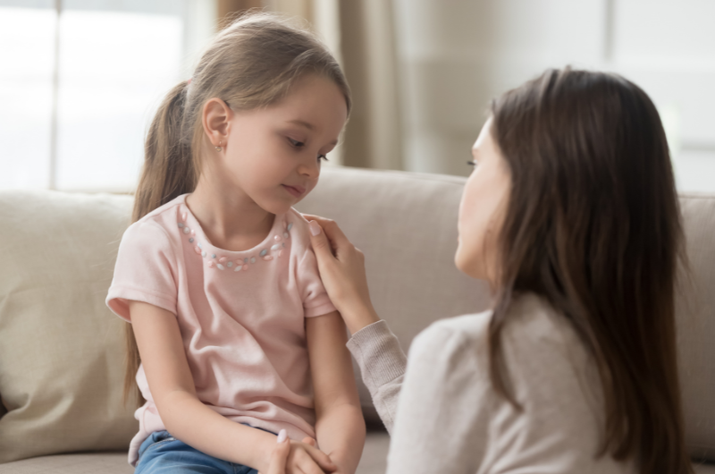 young girl on the couch looking sad with her mom kneeling down next to her taking with her