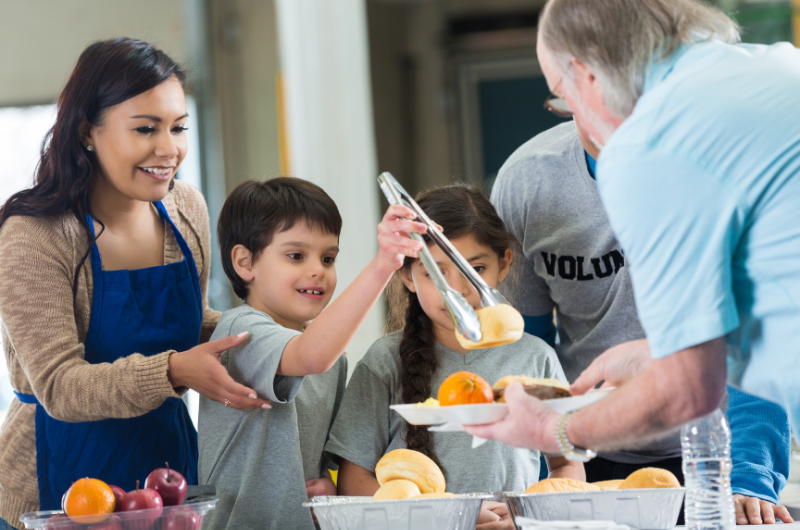 mom with son and daughter volunteering to help serve food at a soup kitchen boy is giving a elderly man a roll